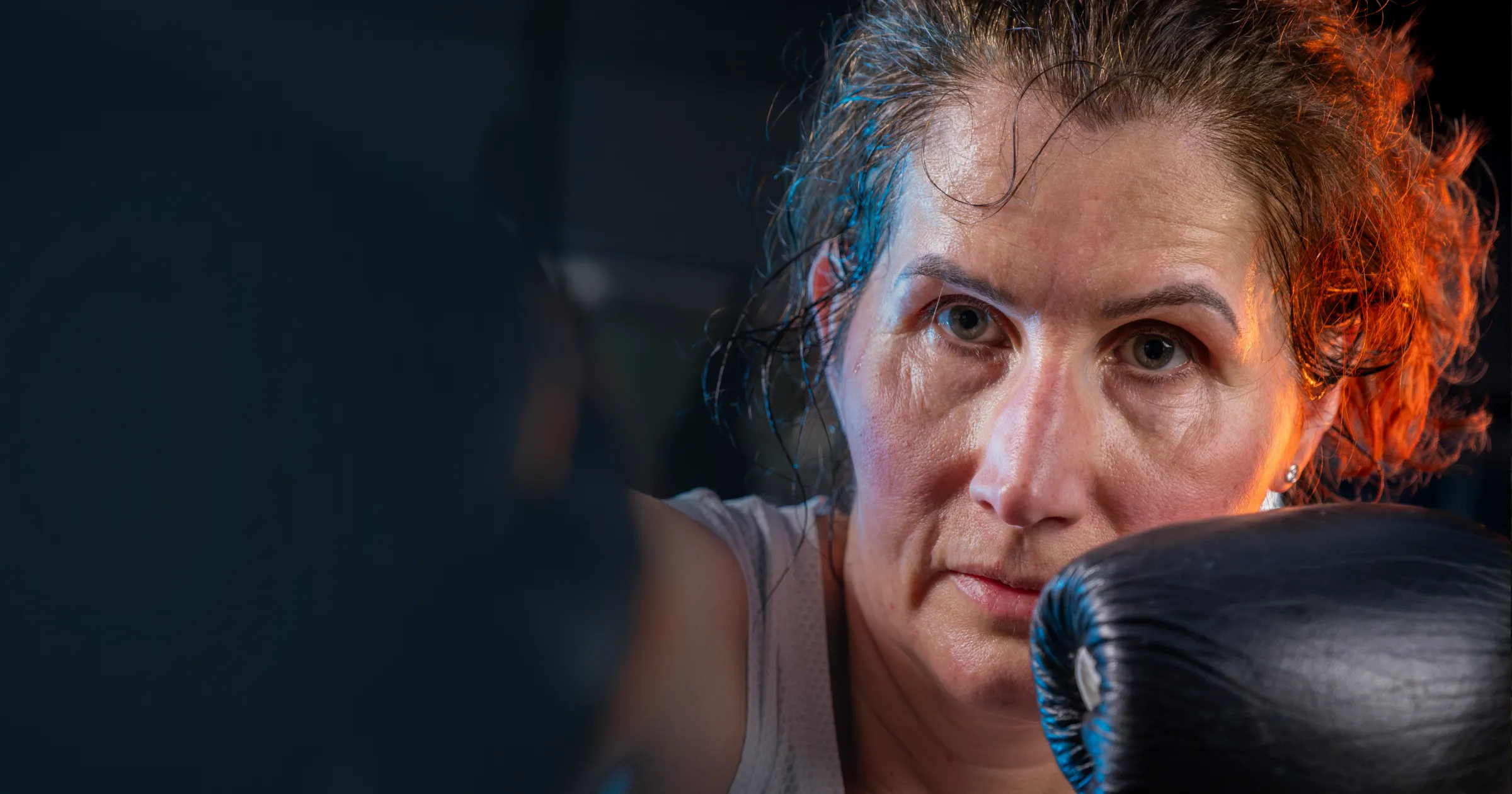 A determined woman with wet hair, wearing boxing gloves, focuses intently during training. Her serious expression conveys strength and concentration.